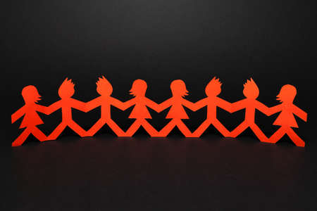 Group of paper doll holding hands. Teamwork concept papercraft. Orange dolls on black wooden backgroundの写真素材