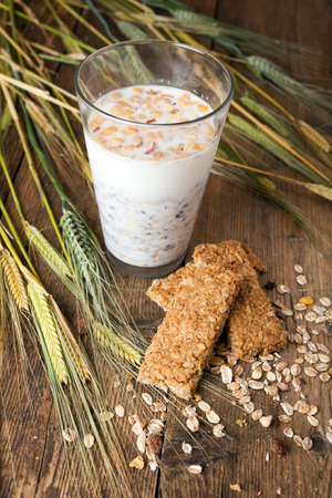 Muesli within a glass of milk, cereal bars and dry wheat on wooden table