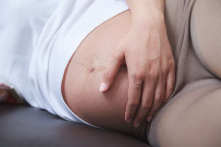 Beautiful pregnant woman holding her hands on bare tummy while sleeping on sofaの写真素材