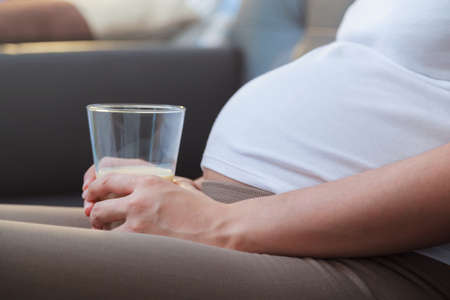 Pregnant woman holding a cup of milk while sitting on sofaの写真素材