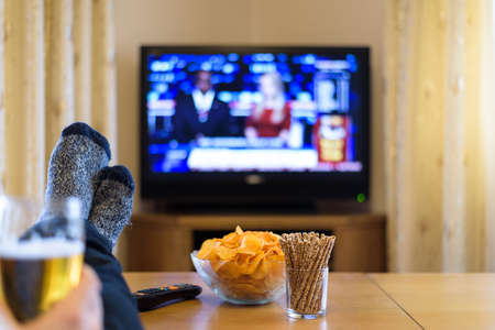 watching TV news with feet on table, eating snacks and drinking beerの写真素材