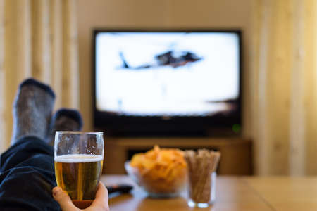 watching TV war movie (military helicopter) with feet on table, eating snacks and drinking beerの写真素材
