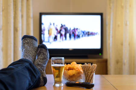 TV, television watching (news about refugees) with feet on table eating snacksの写真素材