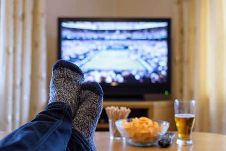 Watching tennis match on TV set (television) with feet on table and eating snacks  stock photoの写真素材
