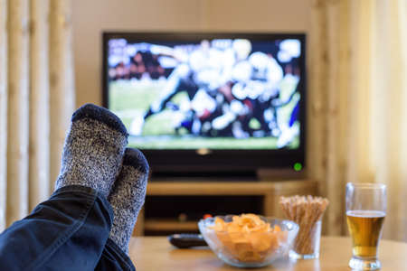 man watching American football match on TV (television) with feet on table, eating snacks - stock photoの写真素材