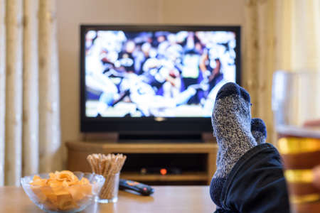 man watching American football match on TV (television) with feet on table, eating snacks - stock photoの写真素材