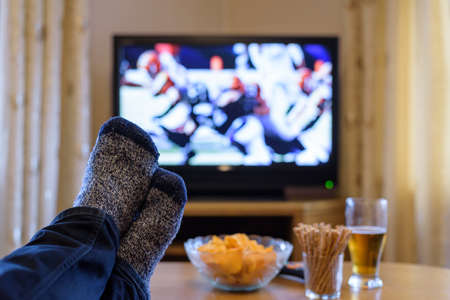 man watching American football match on TV (television) with feet on table, eating snacks - stock photoの写真素材