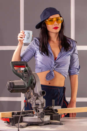 Attractive a happy young woman in blue shirt standing with cup at workbench full of tools and safety equipment in workshop looking at camera and smiling .の写真素材