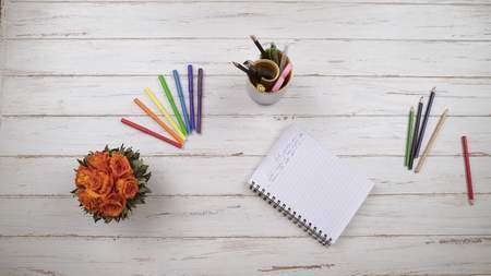 A girl writes a letter to a friend on a white wooden table next to flowers and colored pencils. Top...
