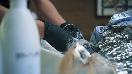 Hairdresser in professional gloves is colouring womans long blonde hair in hair salon above the sink