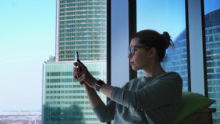 Hipster female in glasses is using apps on her mobile phone and taking photos of skyscrapers view....