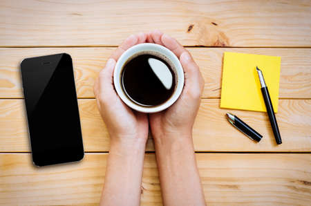hand holding coffee cup with smartphone and notepad on wood table top viewの写真素材