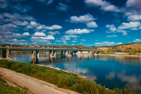 Shore of a broad river with a bridge, reflection of clouds in waterの写真素材