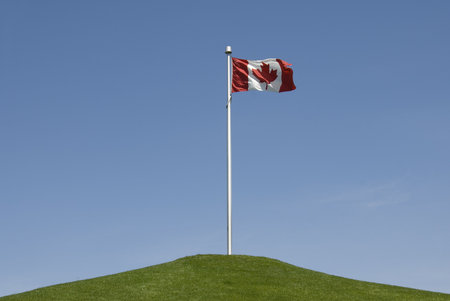 A Canadian flag stands atop a grassy hill against a bright blue sky.の写真素材