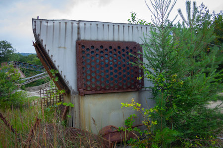 A rusted metal building with a window that is covered in holes. The building is surrounded by trees and grassの写真素材