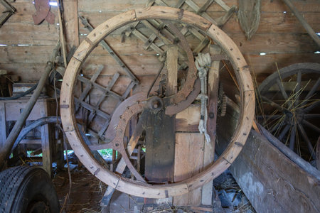 A large wooden wheel with a metal axle in the center. The wheel is old and rusted, and it is a part of a machine. The scene is set in a workshop or a barn, with other old machineryの写真素材