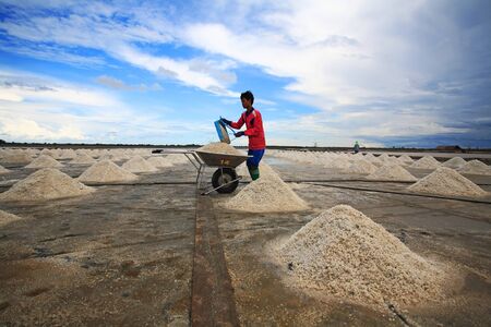 SAMUT SAKHON, THAILAND-APR 16  Unidentified man shovels salt into wheelbarrow at salt pan against blue sky on April 16, 2013 in Samut Sakhon, Thailand の写真素材