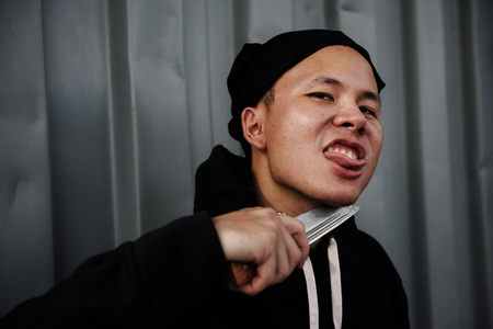 psychosis asian young killer or slayer man with black hoodie jacket holding a knife at his neck and showing tongue to frighten a scared victim. Halloween and murderer concept with copy space for text.の写真素材