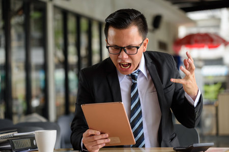 Asian angry businessman wearing glasses, white shirt, black suit looking and yelling at digital...