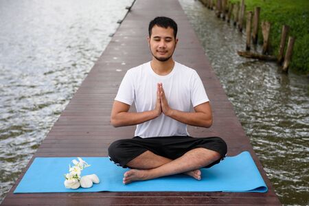 Young Asian smile handsome man, 20-30 years old, Doing Yoga meditation exercise by lotus pose on wooden bridge above peaceful lakes. Healthcare and Fitness conceptの写真素材