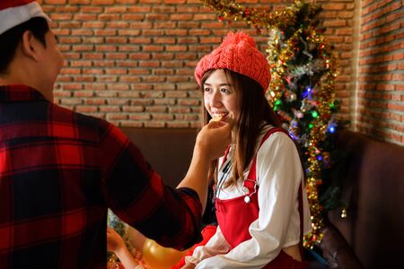 Cute Asian girl looking at boyfriend and eating snack he feed. Lovely happy couple celebrate Christmas or X'mas party, Holiday celebration concept with copy space for text.の写真素材