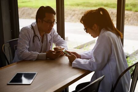 Two happy doctor couple playing smartphone and relaxing at clear wooden table with tablet pc near restaurant window. 4g Technology for working people to explore world. Love in office.の写真素材