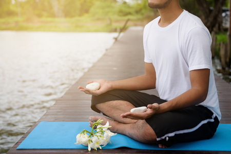 Yoga Male meditation exercise with white rock on hands on wooden bridge at lakes against sunlight. Healthcare and Fitness concept with copy space for text.の写真素材