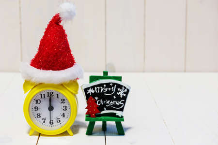 Yellow clock showing midnight time at 12 with red Santa hat and Merry xmas sign on wooden background with copy space for text. Christmas and New year celebration concept for home interior decorationの写真素材