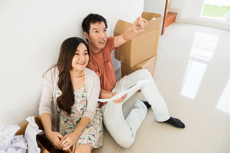 Asian young couple plan the new blank house. They look at a blueprint and happy smile husband point...