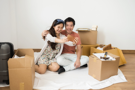 Asian happy couple taking selfie by smartphone and sitting on floor with blueprint, household equipment, luggage, and moving boxes at new home. People lifestyle with technology. start of family life.の写真素材