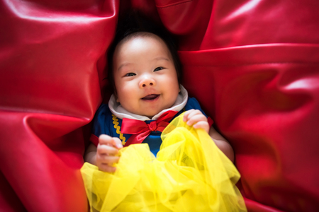 Portrait of happy cute Asian baby, 3 months old with fantasy outfit costume, relax and lie on vibrant red bean bag. Adorable beautiful white girl look at camera.の写真素材