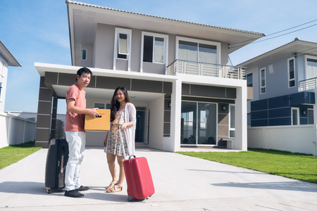 Happy Asian young husband and wife couple carrying box and luggage in front of new home. Portrait of couple moving to modern house. Family lifestyle to start. They bought first homeの写真素材