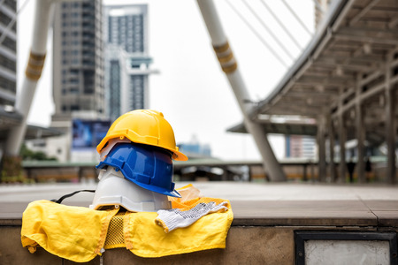 Many colorful Safety helmet hats, gloves, and yellow worker dress on concrete floor with blurred modern city  background. Engineering and construction equipment with copy space  text. Engineer Projectの写真素材
