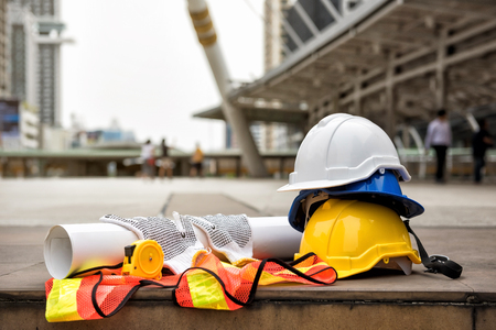 Safety helmet hats, blueprint paper project, measure tape, gloves, and worker dress on concrete floor at modern city with blurred people. Engineer and construction equipment with copy space for text.