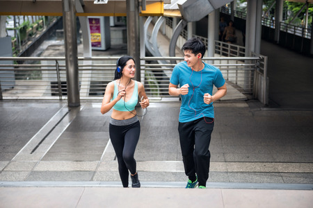 Healthy Asian couple running upstairs at modern city skywalk. Active lifestyle sport people exercising cardio climbing staircase while listenning music from smartphone or mobile phone in town.の写真素材
