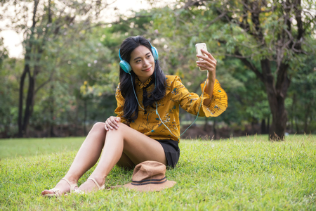 Beautiful Asian Bohemian girl with boho style dress selfie by smartphone or mobile phone in park. Female hipster listening song on meadow garden field. Relax in weekend.の写真素材