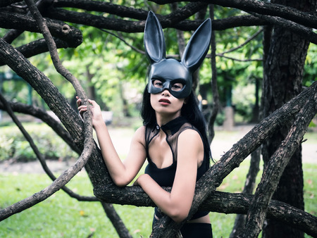 Portrait of beautiful Asian sexy terrify tan woman with fit black dress outfit and rabbit bunny ear mask at old tree in park with copy space for text. Easter holiday with vintage color tone.の写真素材