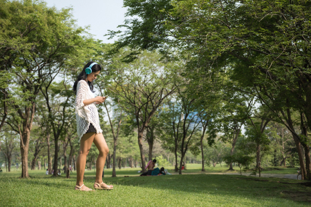 Happy Asian beautiful woman playing smartphone and listening music in summer park. Cute girl walk and relax in garden.の写真素材