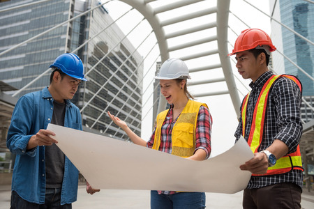 Angry stressed American female boss look at blueprint paper plan and complain Asian engineering man team for project delay. International joint venture of construction site in city.の写真素材