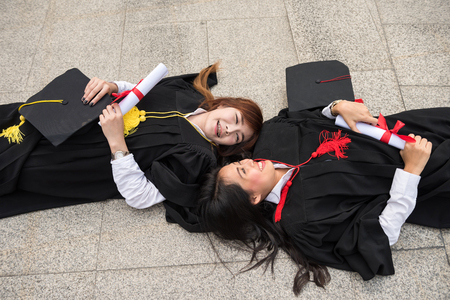 Portrait of Asian beautiful young female students with black graduation gowns hold diploma and lie down on floor. Happy and graceful friendship feeling in commencement day.の写真素材