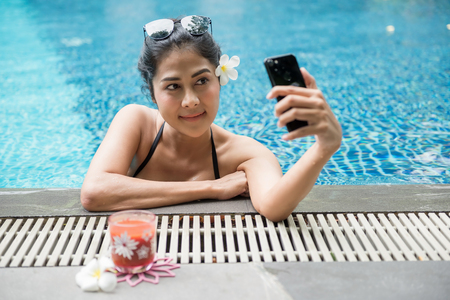 Attractive sexy Asian woman with black two piece swimsuit and sunglasses selfie herself by smartphone nearat swimming pool edge. Beautiful Girl relax during summer vacation.の写真素材