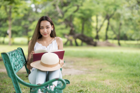 Portrait of Asian beautiful brunette woman read text book and sit on garden chair with greenery tree and nature fresh air. Cute girl in summer park with copy space for text.の写真素材