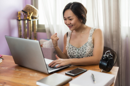 Happy Chinese business woman raise hands after checking good project news from laptop computer using wifi internet from working table in house.の写真素材