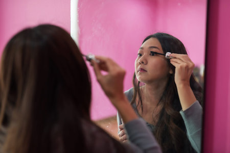 Young Asian woman doing make up of eyelashes in front of mirror in pink room.