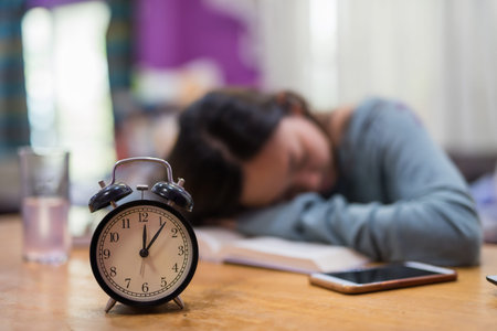 Closeup alarm clock with tired student woman sleep on text book in house. Serious exhauasted Asian...