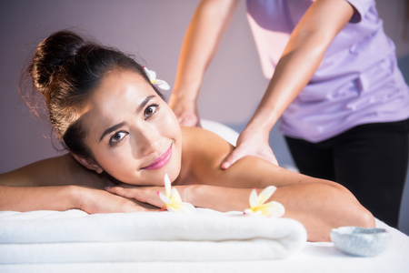 portrait of attractive Asian beautiful woman in spa club. Body care treatment by Thai oil. Cute girl having massage at back by professional massager hands with candle and white Plumeria on bed.の写真素材