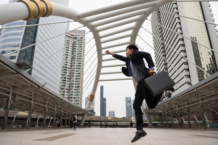 Asian Businessman in formal suit holding bag hurry in modern city. In rush and jump at sky walk of urban town. Business in the city concept. Prevent late of office meeting by running.の写真素材