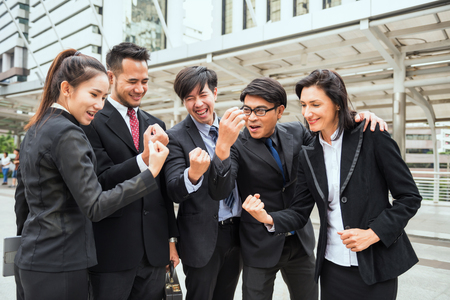 International Business colleague celebrate success with arms raised in modern city after project achievement. Businessman group in suits complete fintech deal.の写真素材