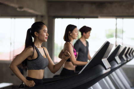 Group of young Asian female and male friends running on treadmills in sport fitness gym. two women...
