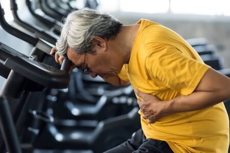 Senior man suffering from chest pain while exercising on biking machine in fitness gym. Old male guy heart attack. Elderly accident from sport training. Healthy lifestyle concept.の写真素材